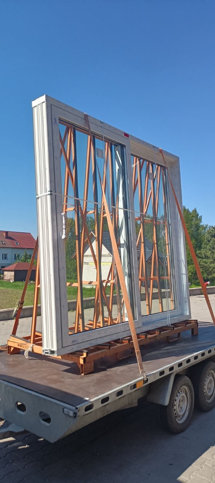 Duże, białe okno przesuwne na przyczepie, zabezpieczone pomarańczowymi pasami transportowymi. W tle budynki i błękitne niebo.