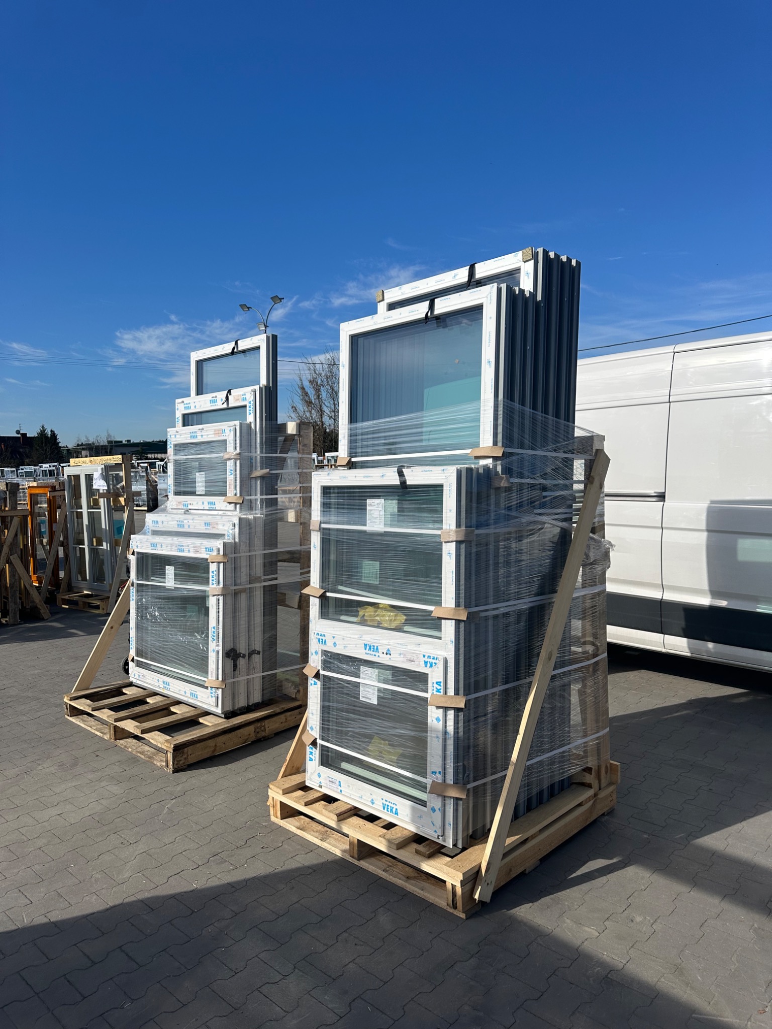 Dwa stosy okien PCV marki VEKA, zabezpieczone folią i umieszczone na drewnianych paletach, gotowe do transportu. Błękitne niebo w tle.