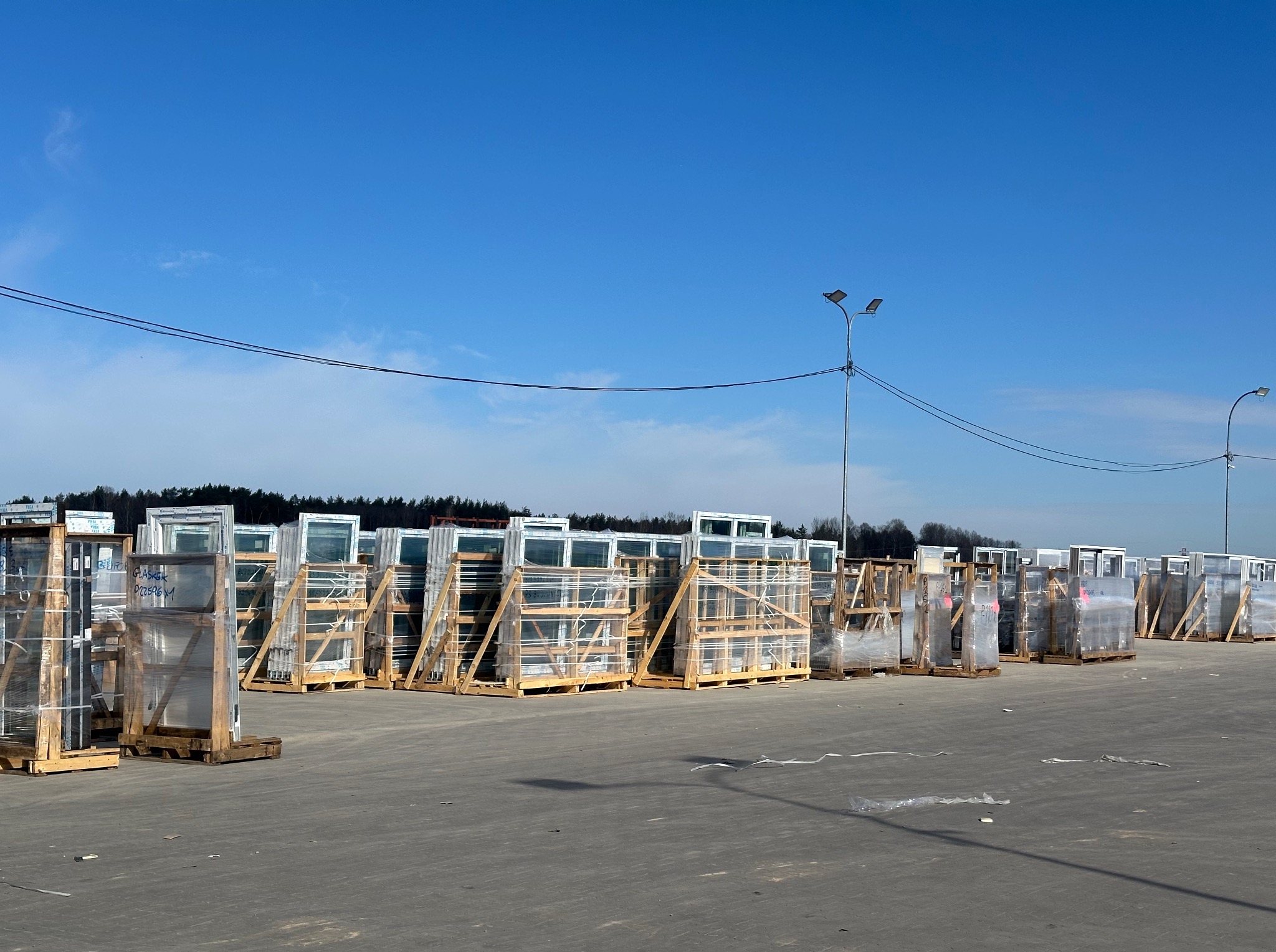 Szeroki kadr: magazyn okiennych ram PCV na paletach, zabezpieczonych folią, pod błękitnym niebem. Widoczne słupy oświetleniowe i fragment lasu w tle.