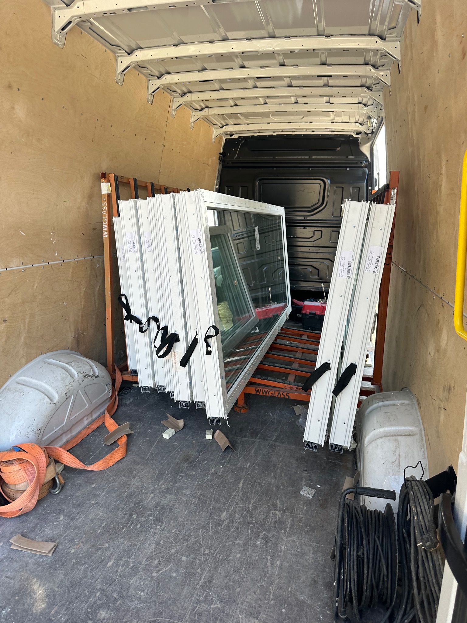 Transport okien PCV w furgonetce, zabezpieczone pasami. Widoczne ramy okienne i listwy przyszybowe na metalowym stojaku. W tle kabina kierowcy.