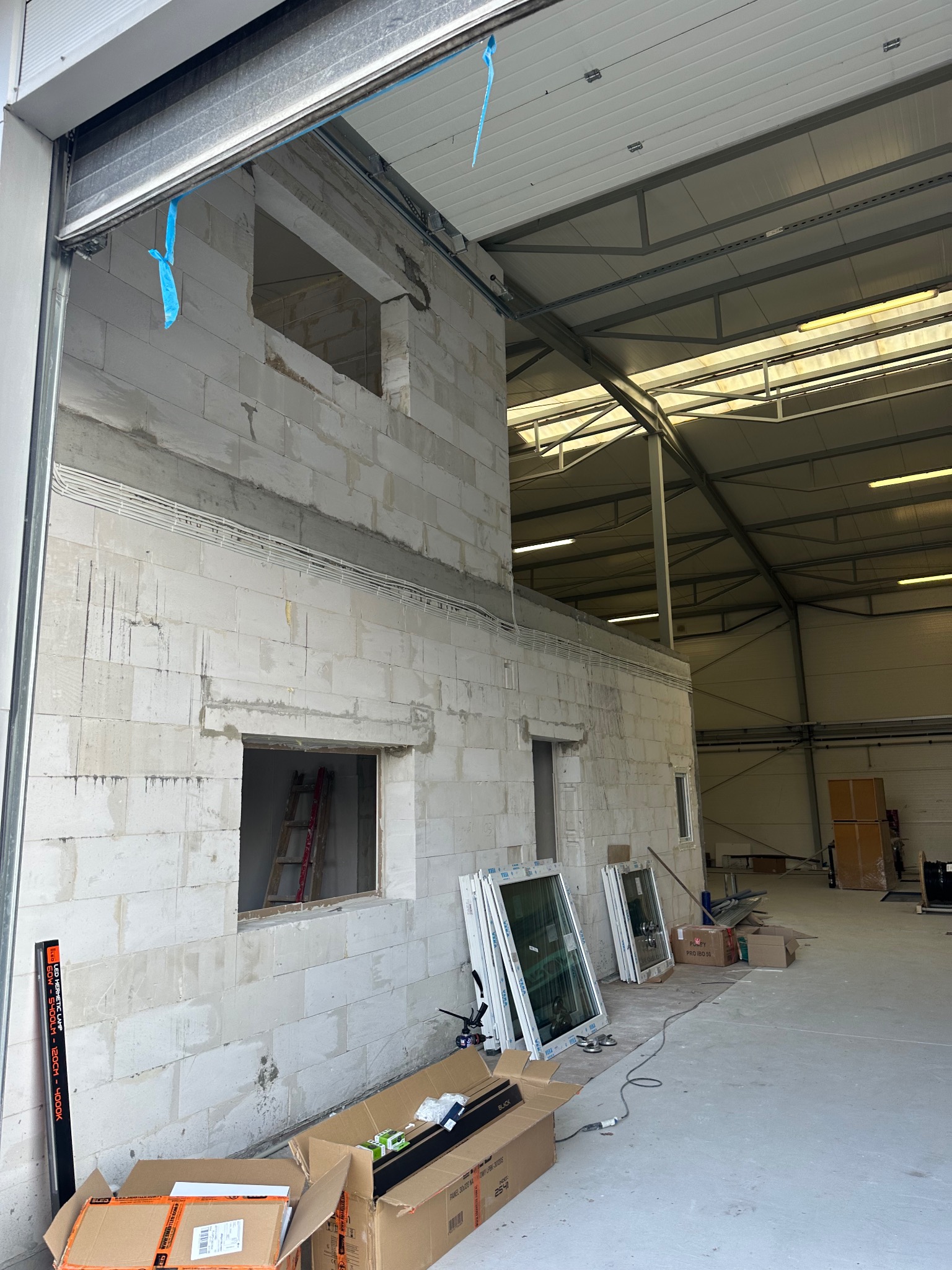 Wnętrze hali w trakcie budowy z widocznymi otworami okiennymi w ścianie z betonu komórkowego. Przy ścianie oparto nowe okna w białych ramach, gotowe do montażu.