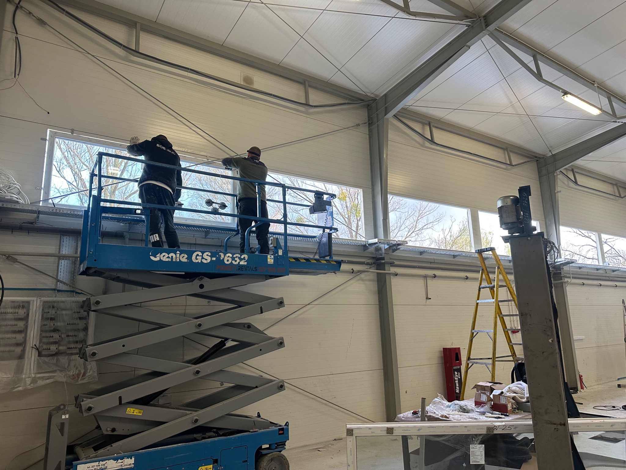Montaż okien w hali produkcyjnej z użyciem podnośnika Genie GS-632. Dwóch monterów pracuje przy oknie, widoczna drabina i narzędzia. Hala z panelową konstrukcją ścian.