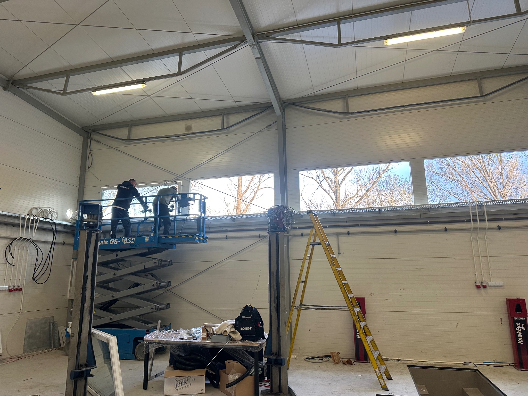 Montaż okien w hali: Dwóch monterów na podnośniku instaluje okno. Widoczna drabina, narzędzia i okno czekające na montaż. Hala z oknami w tle.