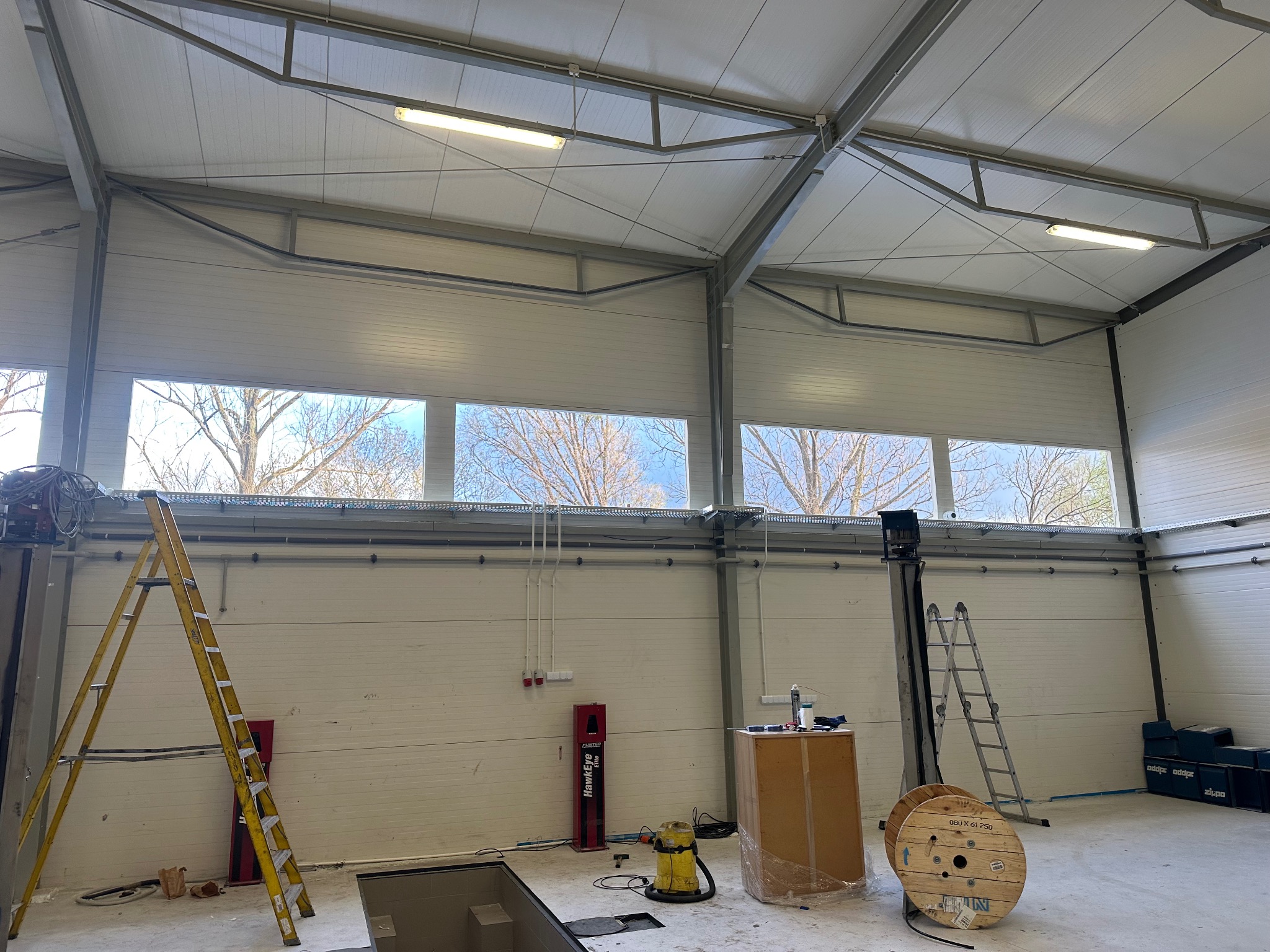 Wnętrze hali z bramą segmentową w Gdańsku. Widoczne okna, drabiny, podnośnik samochodowy i inne wyposażenie. Hala oświetlona lampami jarzeniowymi.