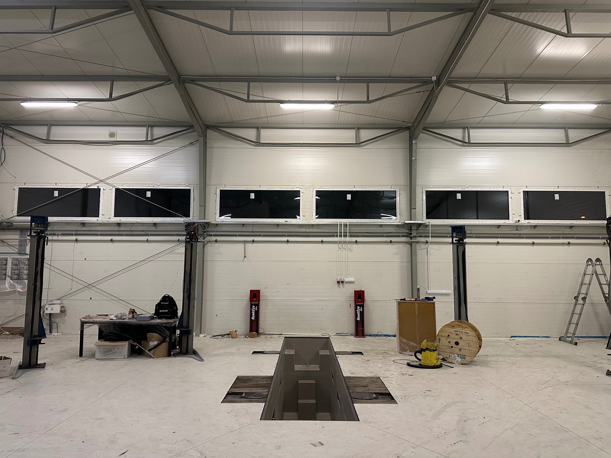 Wnętrze hali warsztatowej z podnośnikami samochodowymi, kanałem rewizyjnym i oknami w górnej części ściany. Widoczne narzędzia i akcesoria warsztatowe.