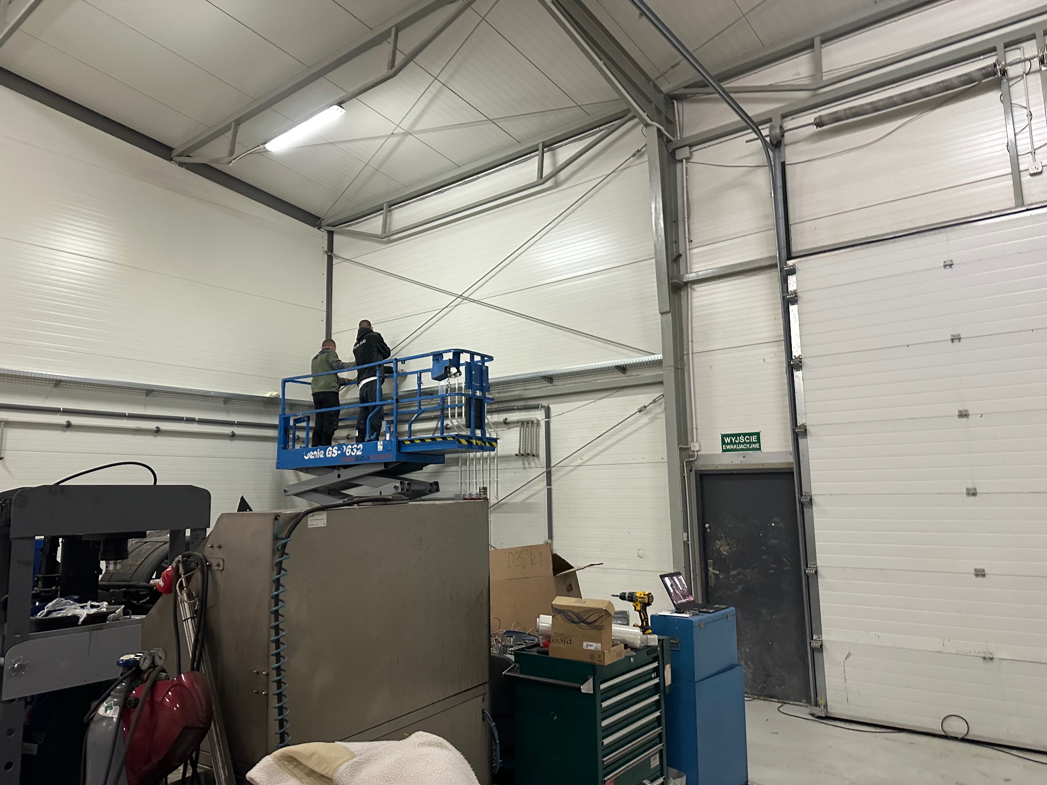 Montaż bramy garażowej segmentowej w hali produkcyjnej. Dwóch monterów na podnośniku koszowym przy suficie. Widoczne narzędzia i elementy konstrukcyjne bramy.