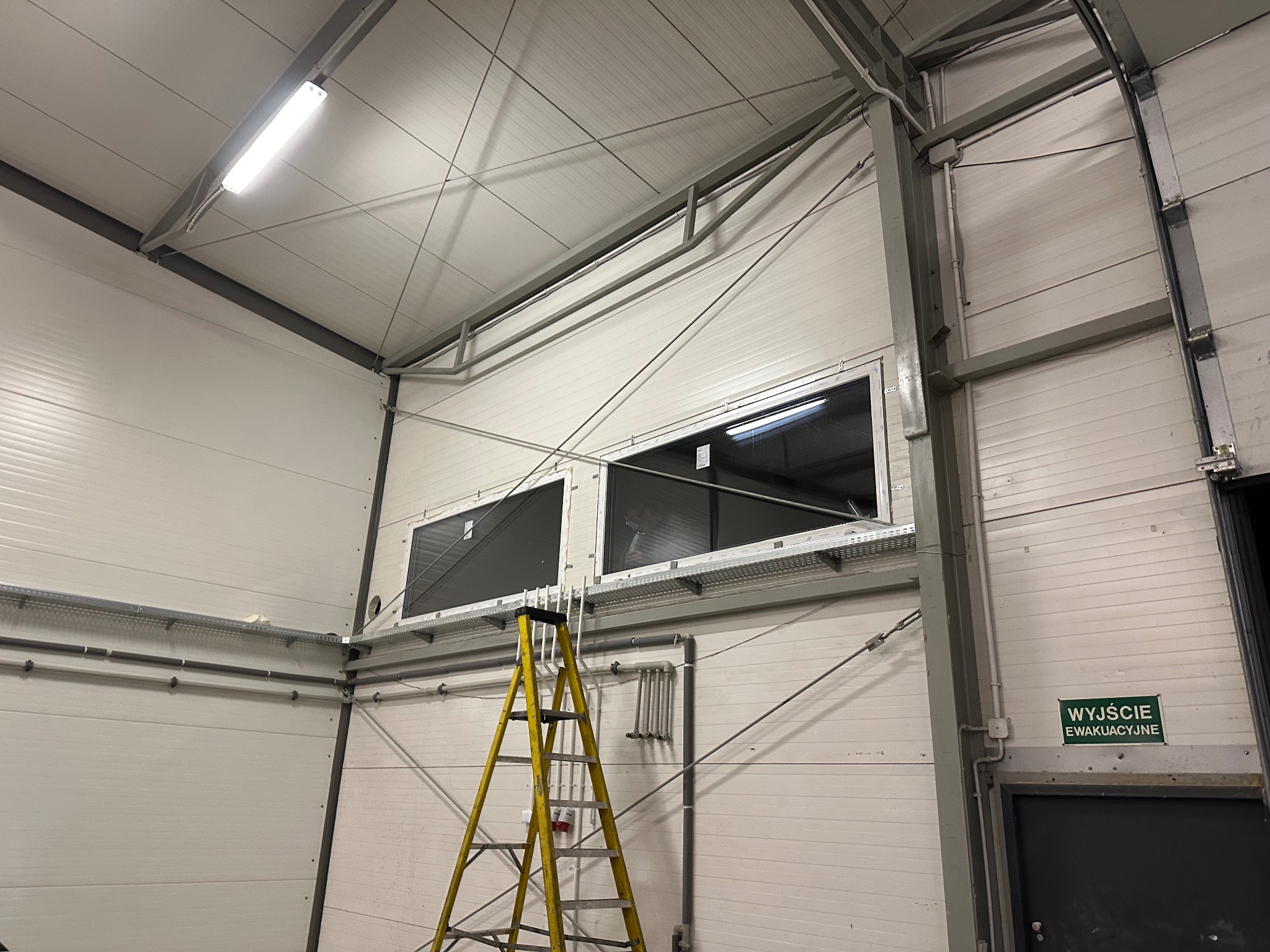 Dwa okna zamontowane wysoko w hali przemysłowej, widoczne z żółtej drabiny. Białe ściany z panelami, metalowa konstrukcja dachu i instalacje. Tabliczka 'Wyjście ewakuacyjne'.