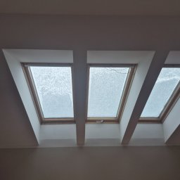 Patryk Puchalski - Trzy okna dachowe w skośnym suficie, oddzielone białymi belkami. Widok na zewnątrz zasłonięty przez śnieg. Minimalistyczne wykończenie wnętrza.