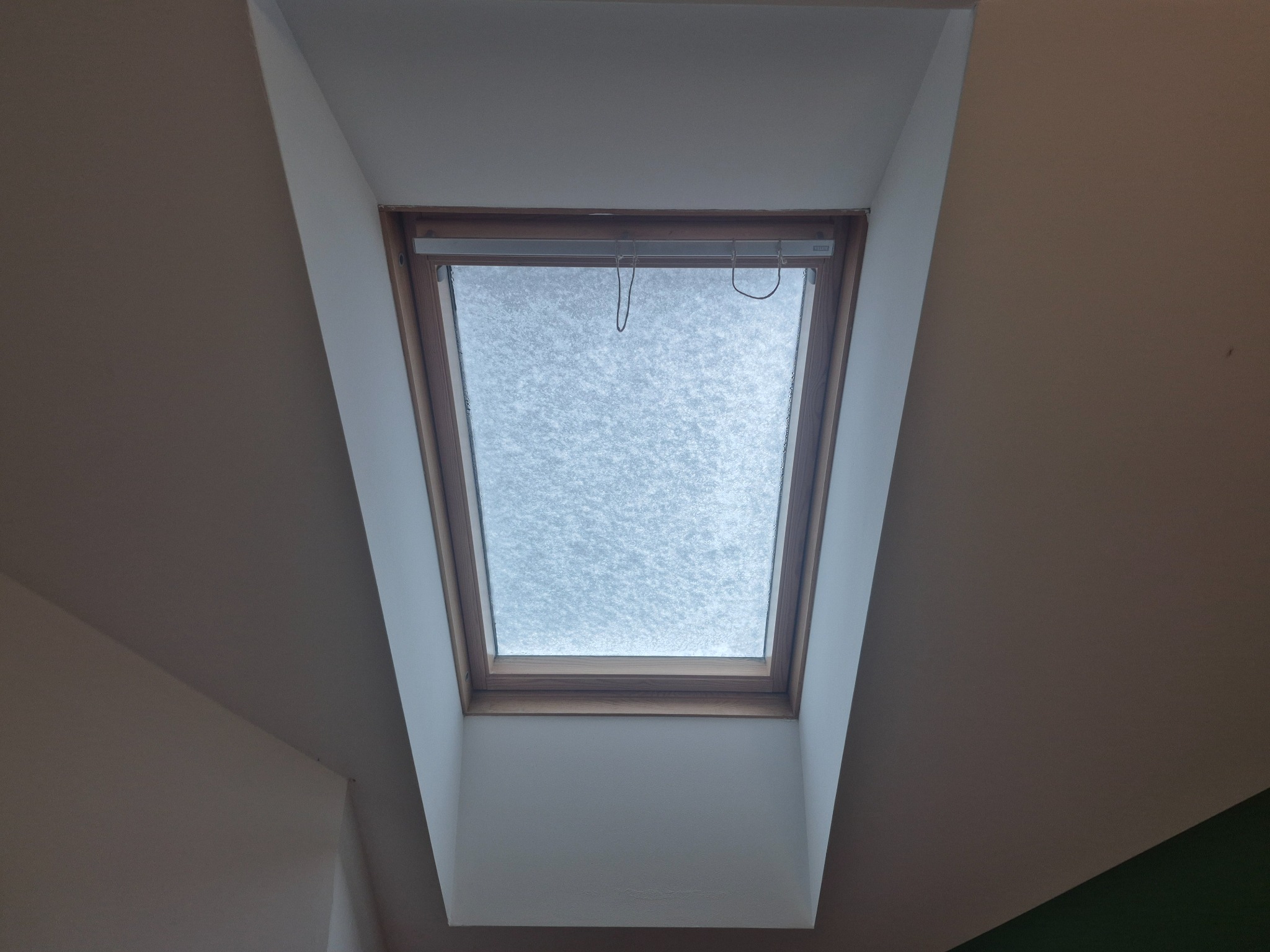 Okno dachowe z drewnianą ramą, widok od dołu, w tle białe ściany poddasza. Widoczna roleta okienna. Ujęcie centralne, symetryczne.