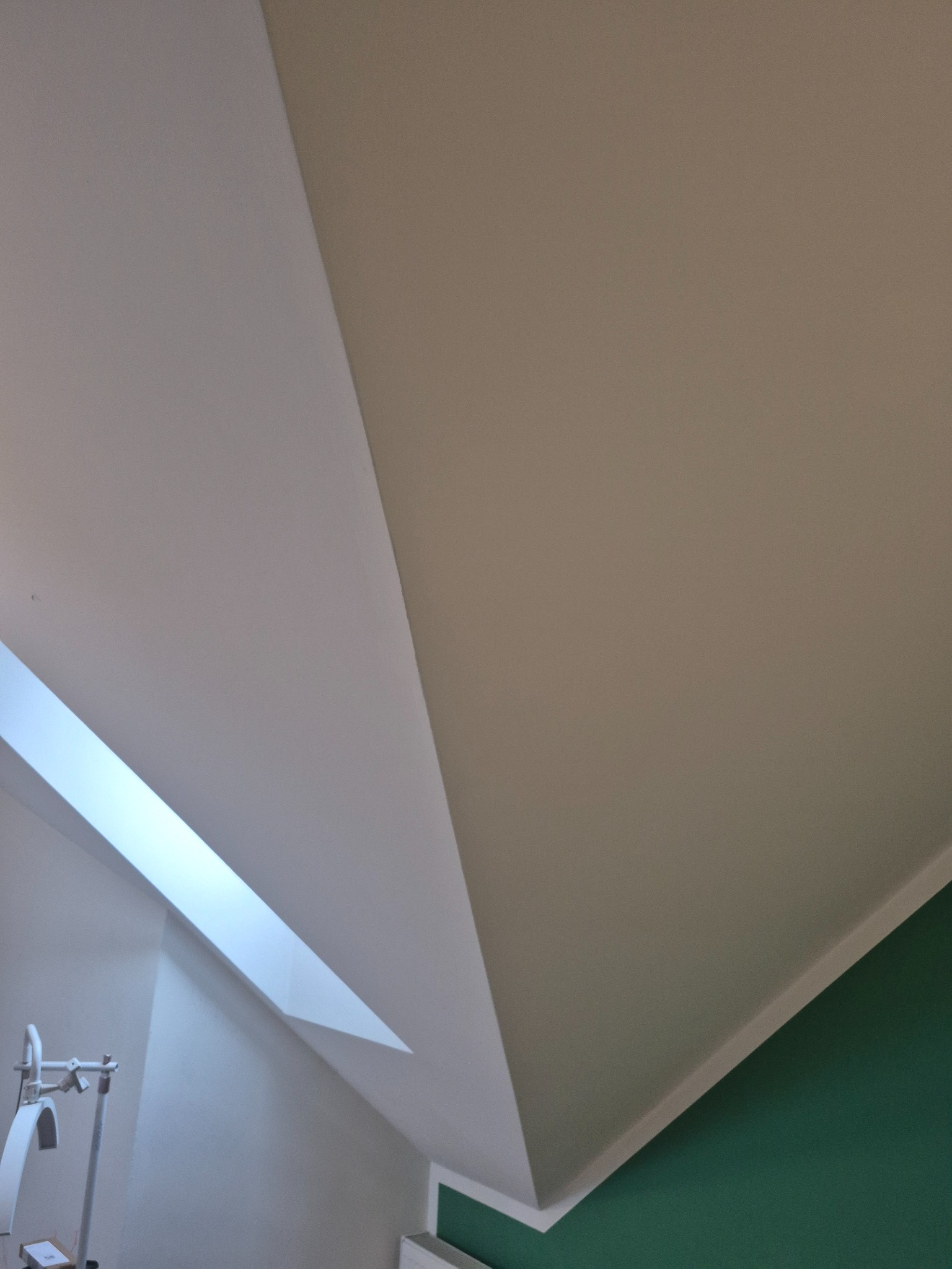 Wykończone poddasze ze skośnym sufitem i oknem dachowym. Połączenie białej i beżowej powierzchni sufitu z zieloną ścianą. Widoczny fragment lampy.