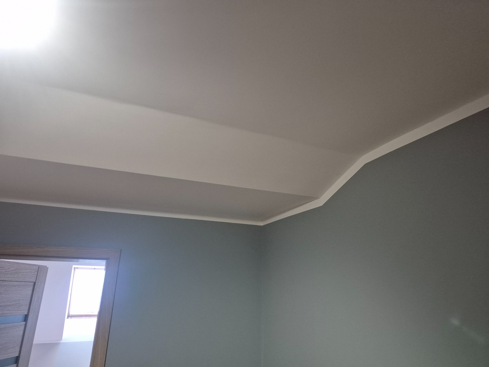 Wykończony sufit podwieszany z listwą dekoracyjną w rogu pokoju o szarych ścianach. Widoczny fragment ościeżnicy drzwiowej.