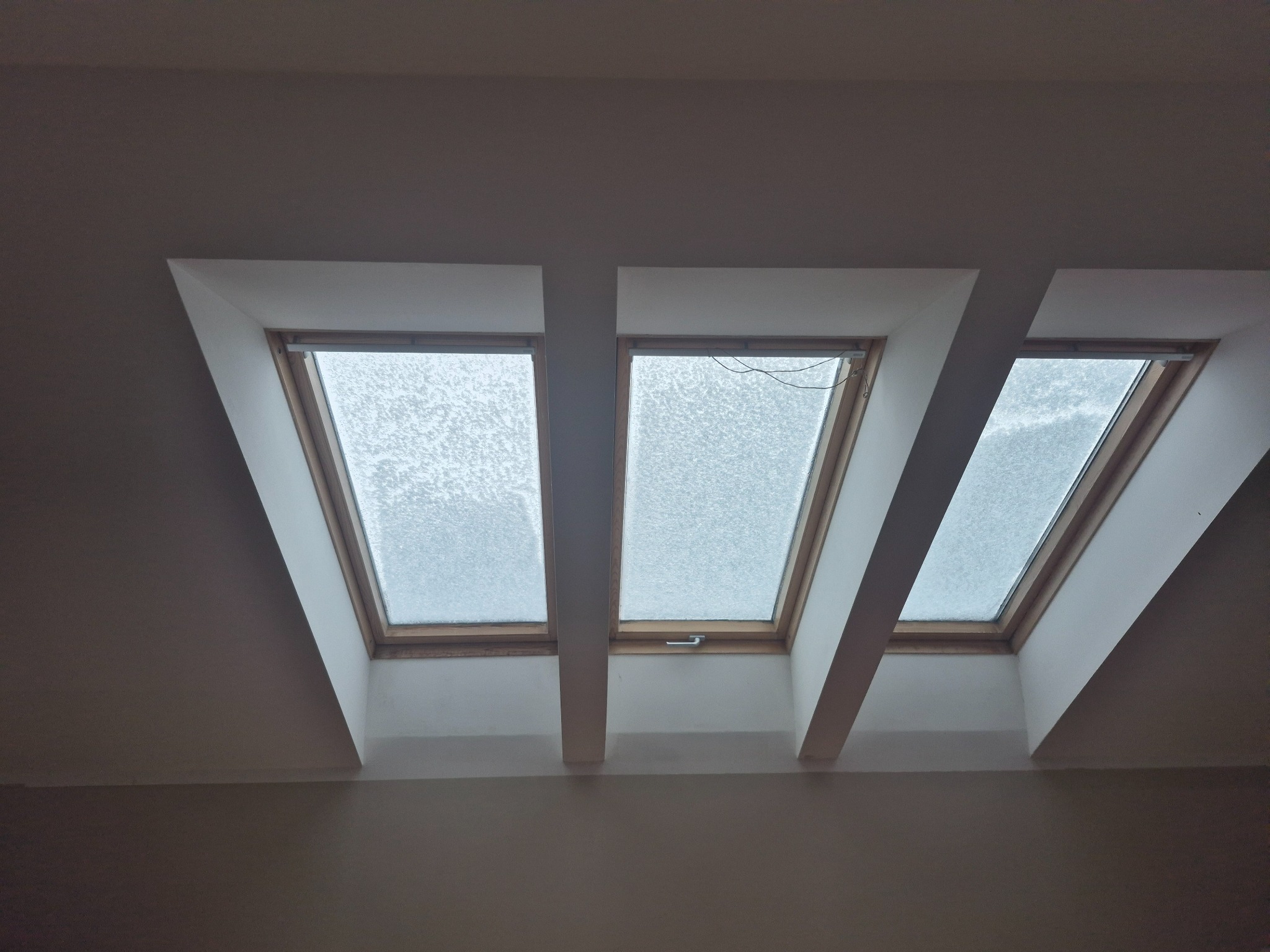 Trzy okna dachowe w skośnym suficie, oddzielone białymi belkami. Widok na zewnątrz zasłonięty przez śnieg. Minimalistyczne wykończenie wnętrza.