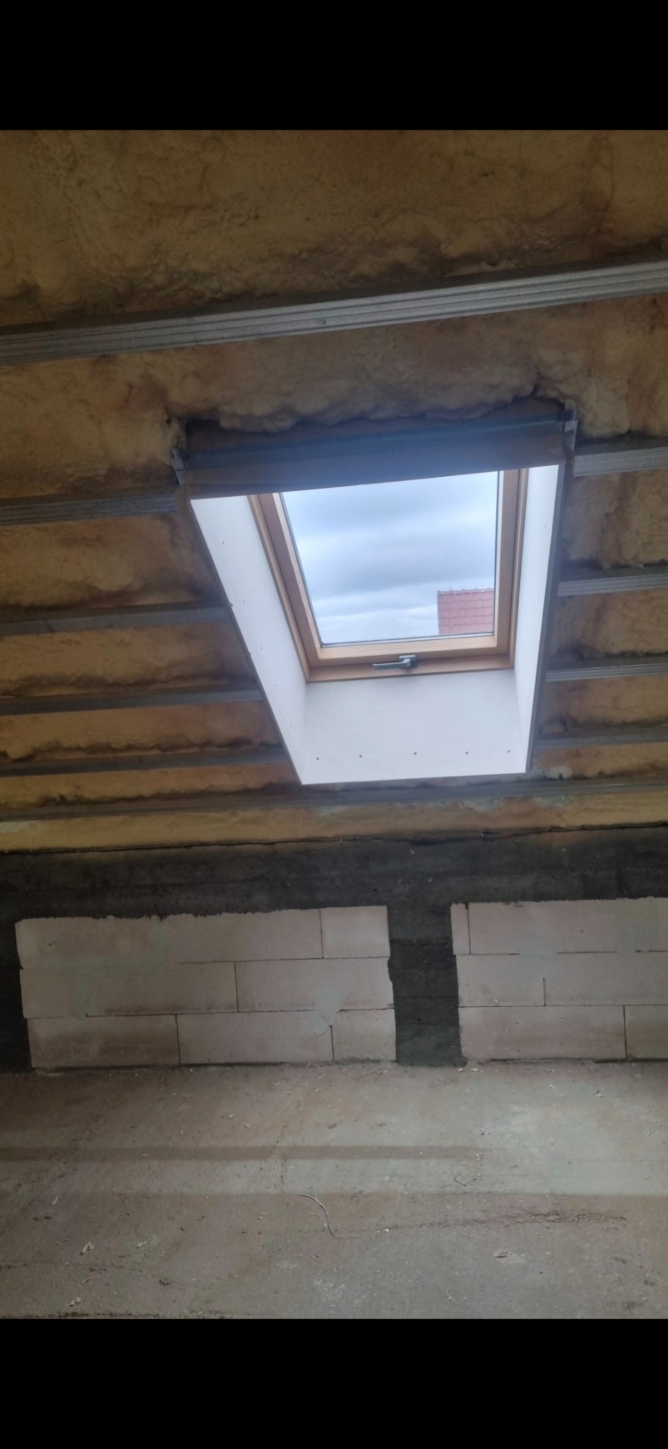 Montaż okna dachowego w surowym wnętrzu: izolacja wełną mineralną, metalowe profile, częściowo wymurowana ściana z bloczków. Widok z dołu na okno i niebo.