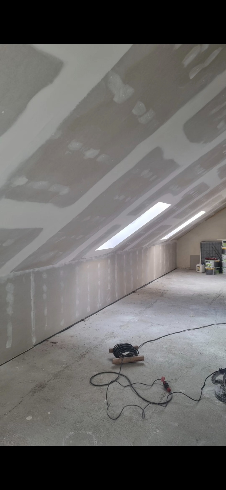 Poddasze po montażu płyt. Widoczne łączenia wymagające szpachlowania, okna dachowe i przewody narzędzi na podłodze. Prace wykończeniowe w trakcie.