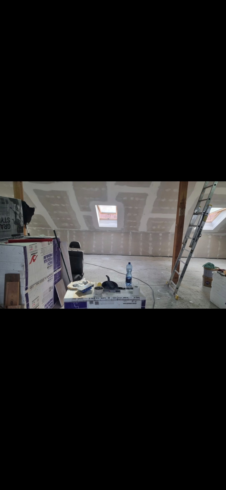 Montaż płyt na poddaszu z oknami dachowymi. Widoczne narzędzia pracy i materiały budowlane. Prace wykończeniowe w trakcie realizacji.