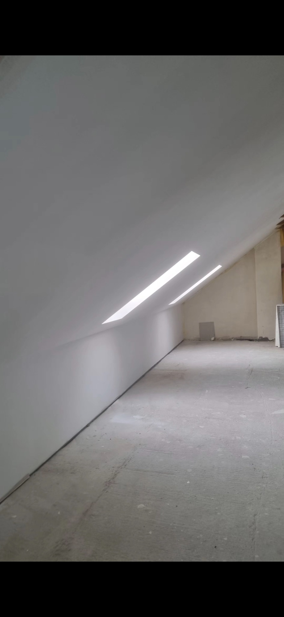 Wykończone poddasze ze ścianami i skosami pokrytymi gładzią, dwa okna dachowe w skosie, surowa podłoga. Widok na fragment pomieszczenia w neutralnej tonacji.