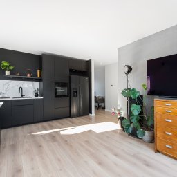 Freedom - Nowoczesna kuchnia z czarnymi szafkami i marmurowym blatem, jasna drewniana podłoga, duży telewizor na komodzie, roślina doniczkowa. Minimalistyczny design.