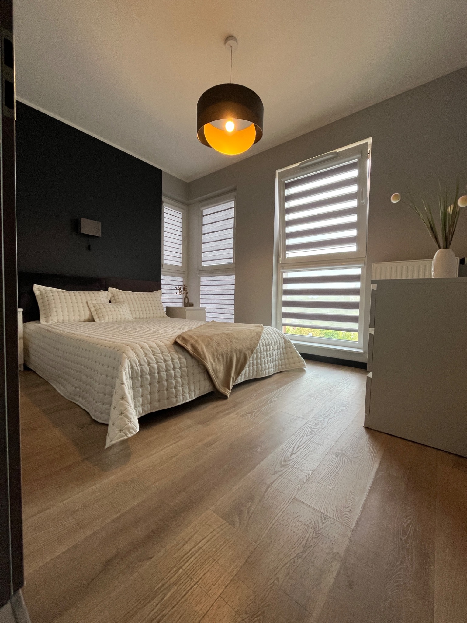 Jasna sypialnia z drewnianą podłogą, dużym łóżkiem z beżową narzutą i oknami z roletami dzień-noc. Czarna ściana za wezgłowiem. Nowoczesny design.