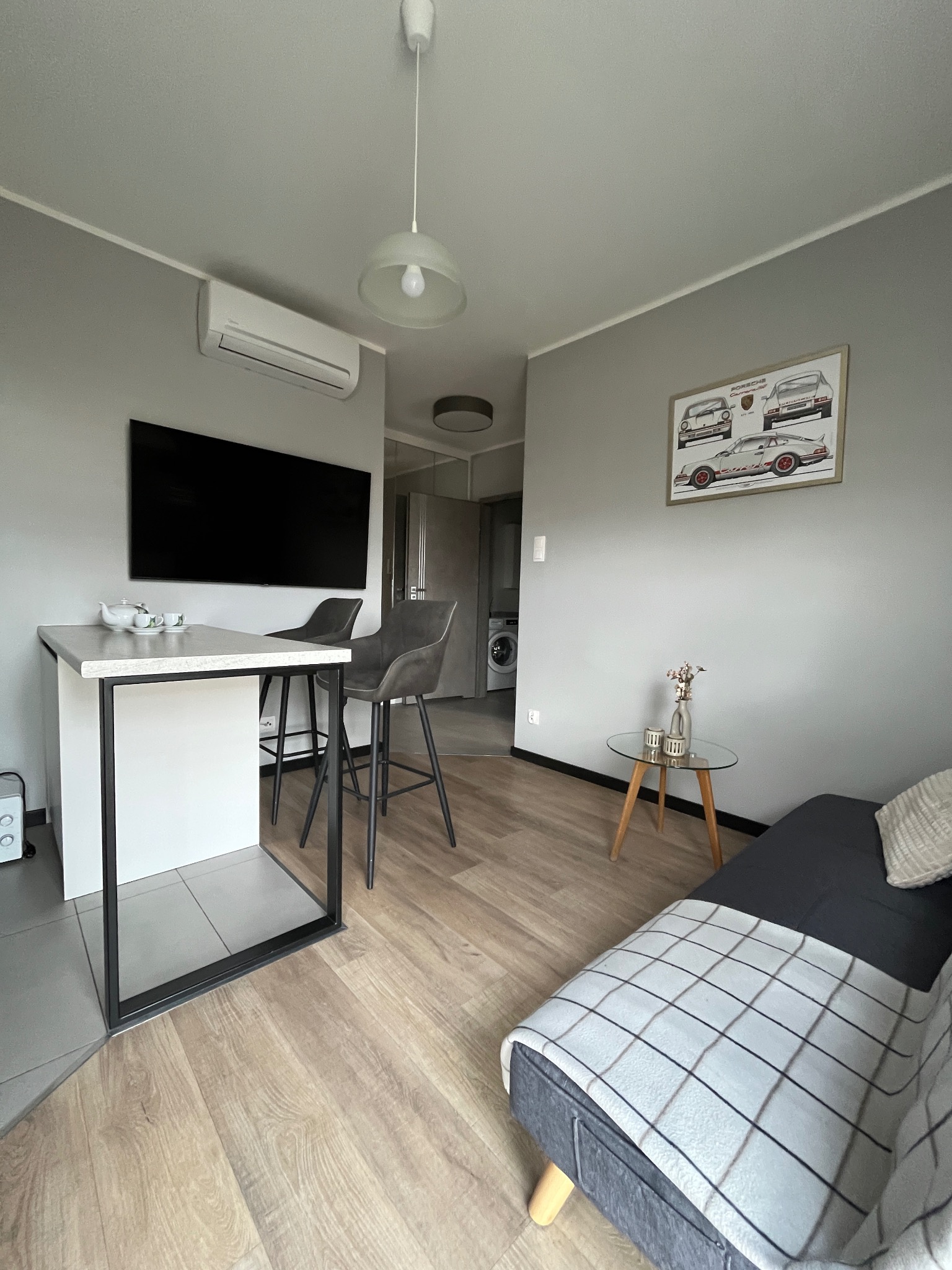 Jasne wnętrze mieszkania z aneksem kuchennym, wysokim stołem barowym i sofą z pledem w kratę. Na ścianie plakat z samochodami Porsche.