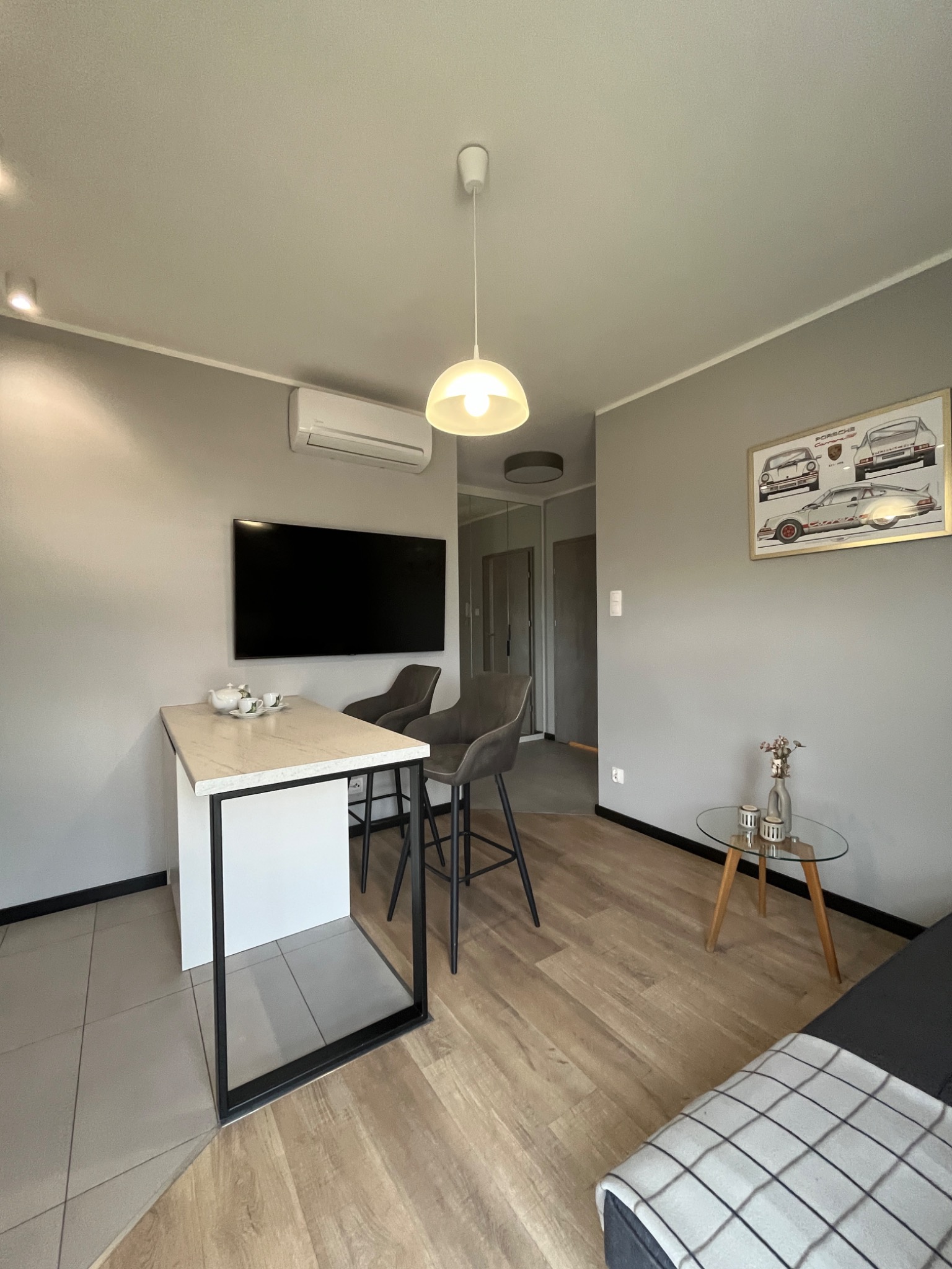 Jasne wnętrze mieszkania z aneksem kuchennym, stołem barowym i telewizorem. Na ścianie plakat z Porsche. Minimalistyczny design, neutralna kolorystyka.