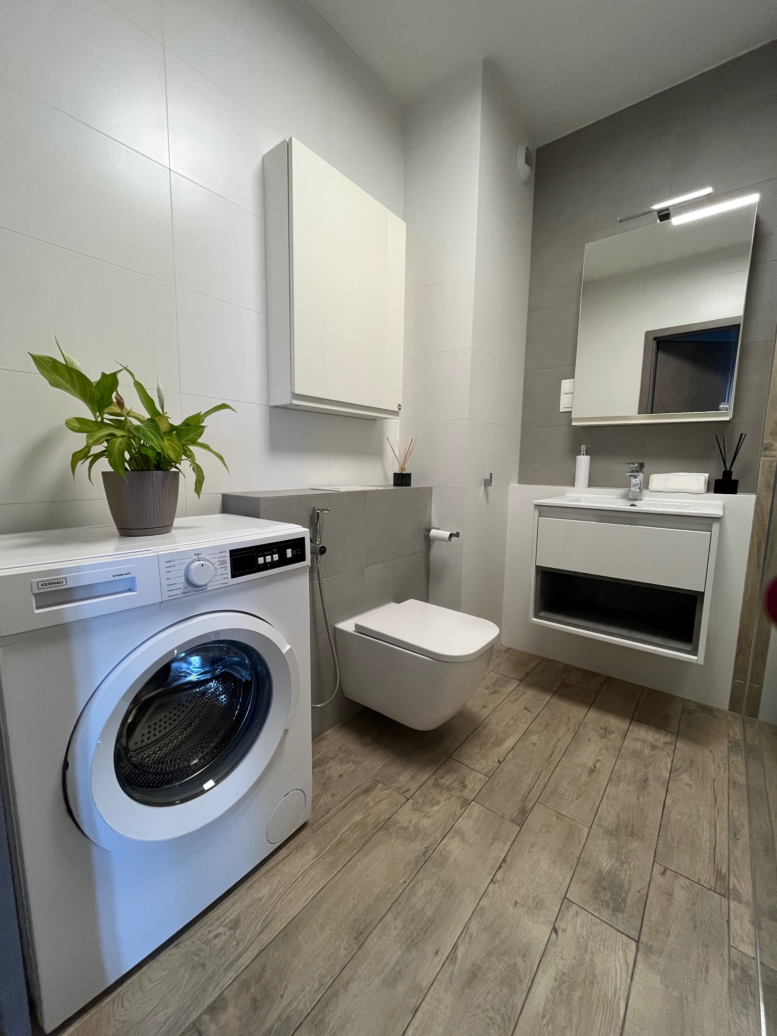 Jasna łazienka z drewnopodobną podłogą, białą pralką Kernau i wiszącą szafką. Na pralce stoi doniczka z zieloną rośliną. Minimalistyczny design.