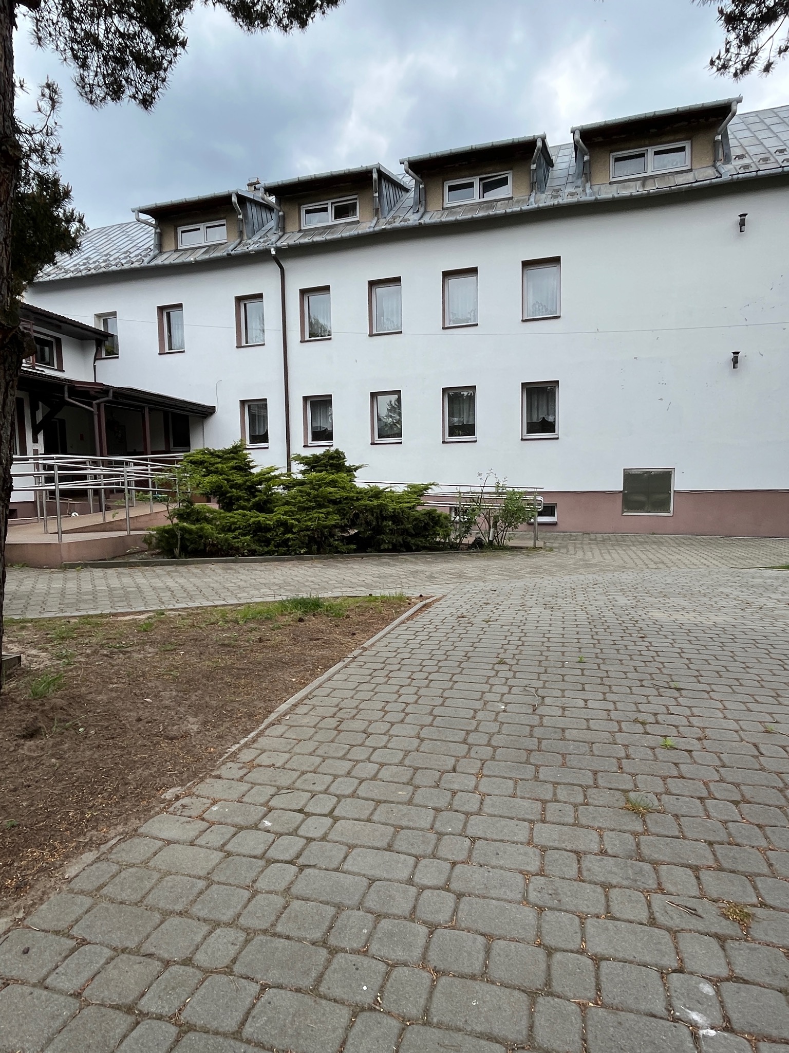 Biały budynek z szarym dachem i oknami z brązowymi ramami, otoczony zielenią i brukowanym chodnikiem. Widoczne poddasze z oknami dachowymi.