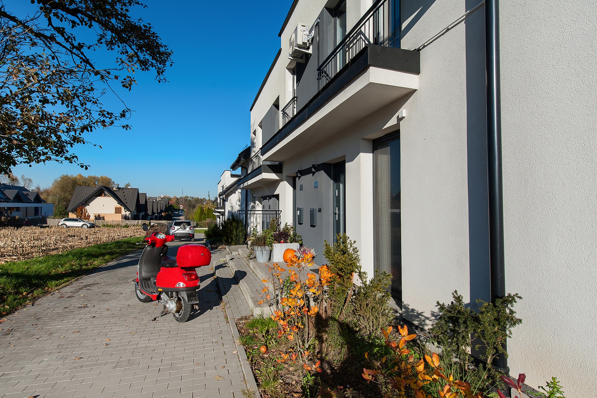 Nowoczesne szeregówki z balkonami, czerwony skuter na chodniku, jesienne dekoracje przed wejściem. Słoneczny dzień, błękitne niebo.