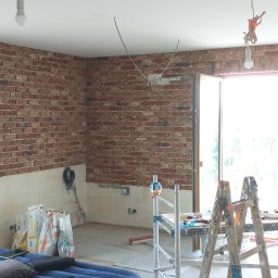 Andrzej Górnis - Wnętrze w trakcie prac wykończeniowych: ściany z cegły klinkierowej do połowy wysokości, worki z zaprawą, drabina malarska. Widoczne okna z niebieskimi ramami.