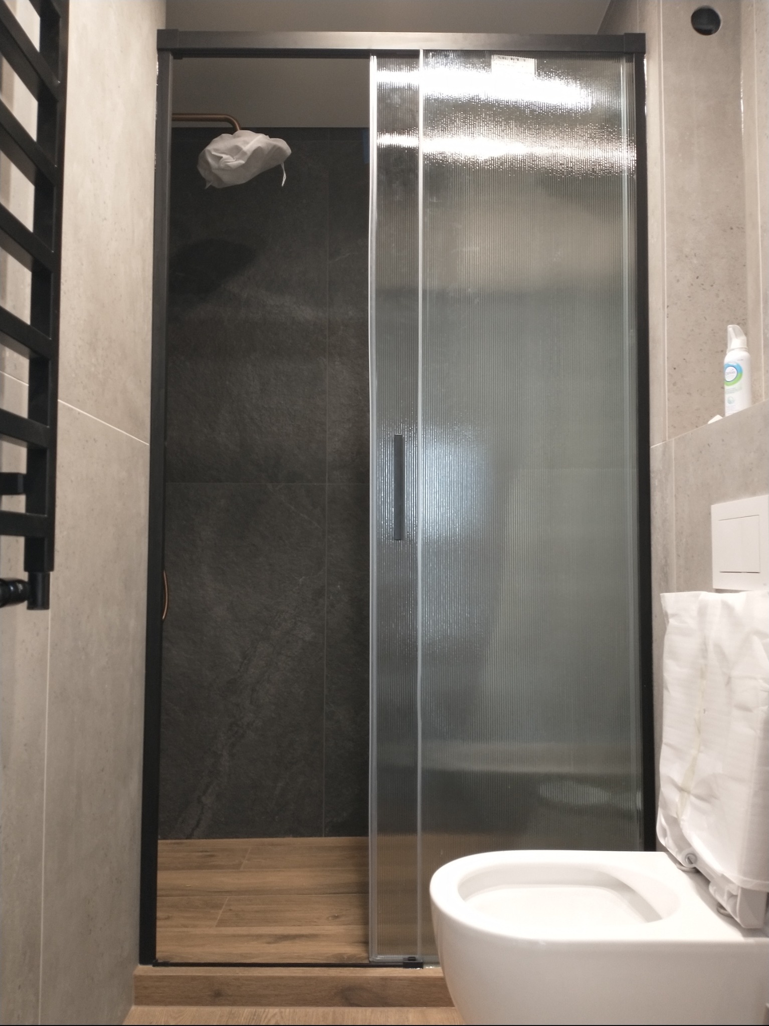 Nowoczesna kabina prysznicowa z przesuwnymi drzwiami z prążkowanego szkła w łazience wykończonej szarymi płytkami. Widoczny fragment toalety i grzejnika.