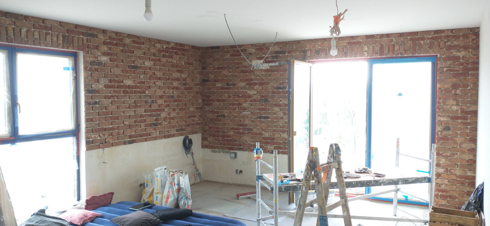 Wnętrze w trakcie prac wykończeniowych: ściany z cegły klinkierowej do połowy wysokości, worki z zaprawą, drabina malarska. Widoczne okna z niebieskimi ramami.