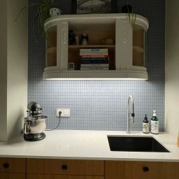 NEUROOM Katarzyna Nojman - Kuchenna aranżacja w Wejherowie: biała szafka wisząca nad blatem z czarnym zlewem, mozaika na ścianie i mikser stojący. Nowoczesny design z elementami retro.