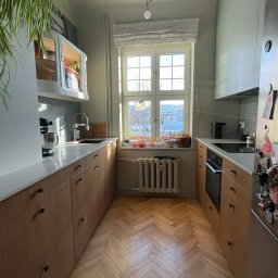 NEUROOM Katarzyna Nojman - Jasna kuchnia z drewnianymi szafkami, parkietem w jodełkę i dużym oknem z widokiem na miasto. Na blacie mikser i owoce, a na lodówce magnesy.