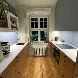 NEUROOM Katarzyna Nojman - Ustawna kuchnia z drewnianą podłogą w jodełkę, jasnymi blatami i niebieską mozaiką na ścianie. Okno z roletą rzymską i roślinami dodaje przytulności.