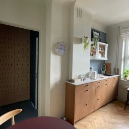 NEUROOM Katarzyna Nojman - Jasna kuchnia z drewnianymi szafkami, parkietem w jodełkę i okrągłym stołem. Zegar na ścianie. Minimalistyczny design z roślinami doniczkowymi.