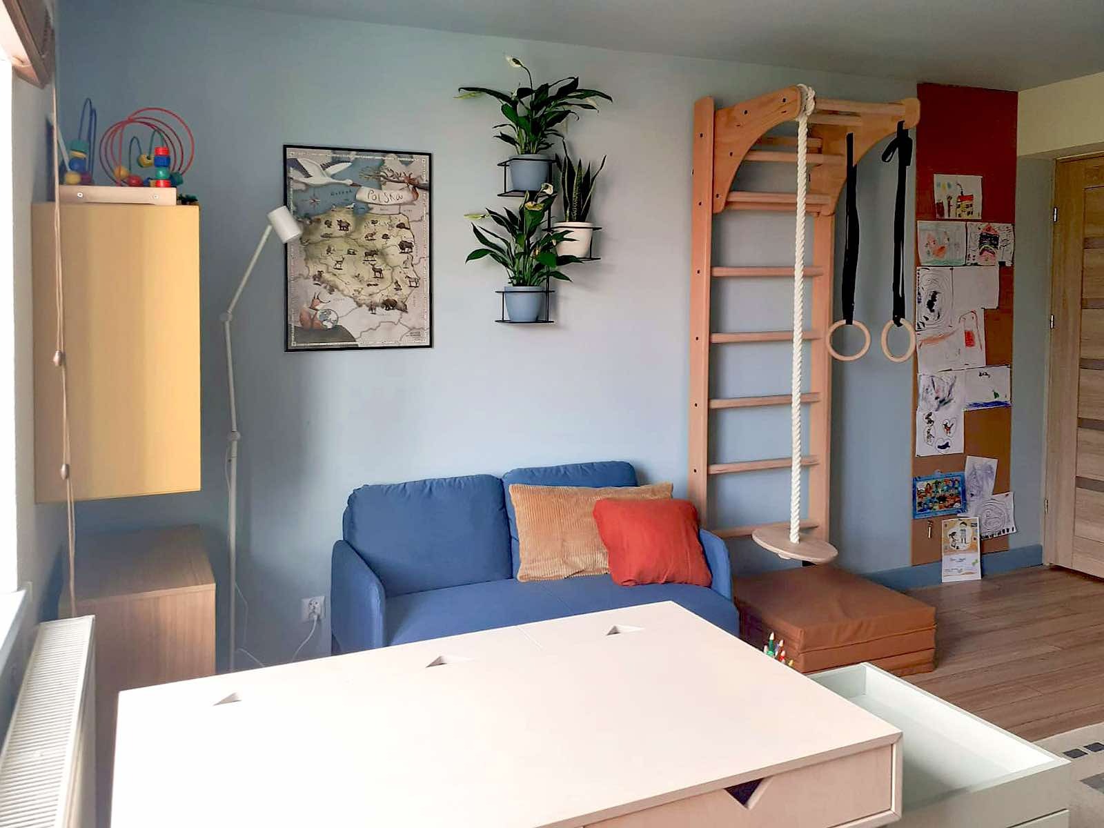 Pokój dziecięcy z niebieską sofą, drewnianą drabinką gimnastyczną i biurkiem z wysuwaną szufladą. Na ścianie mapa Polski i rośliny w doniczkach.