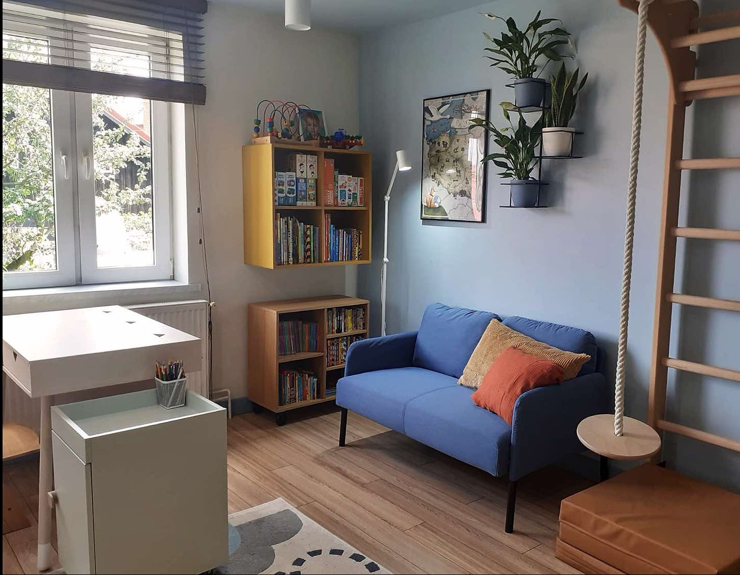 Pokój dziecięcy z niebieską sofą, drewnianą drabinką gimnastyczną i biurkiem przy oknie. Na ścianie półki z książkami i roślinami. Jasne drewno i pastelowe kolory.