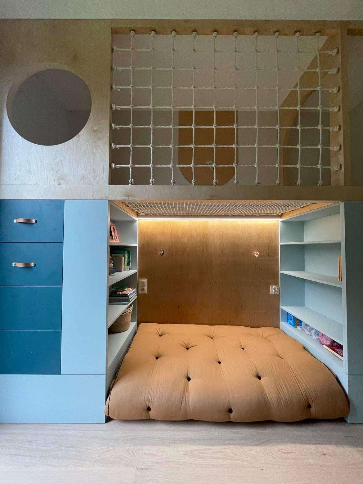 Kreatywna aranżacja pokoju dziecięcego: łóżko-antresola z siatką do zabawy, niebieskie szafki z drewnianymi uchwytami i półki na książki. Oświetlenie LED nad łóżkiem.