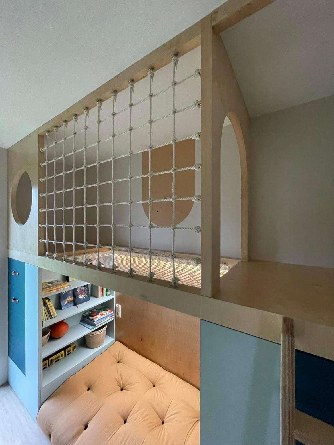 Kreatywny pokój dziecięcy z antresolą zabezpieczoną siatką sznurkową, zabudowany regał z książkami i materacem. Nietypowe rozwiązanie w jasnych barwach.