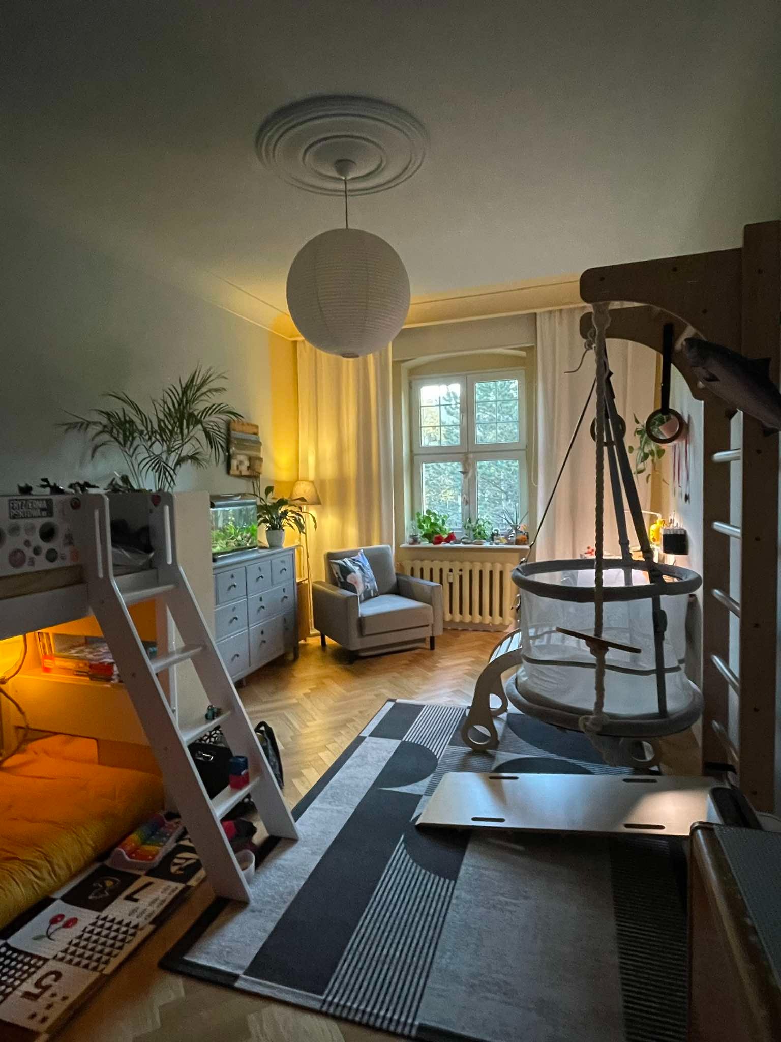 Pokój dziecięcy z łóżkiem piętrowym, komodą z akwarium, fotelem i wiszącym łóżeczkiem. Parkiet, dywan w geometryczne wzory, okno z roślinami. Ciepłe oświetlenie.