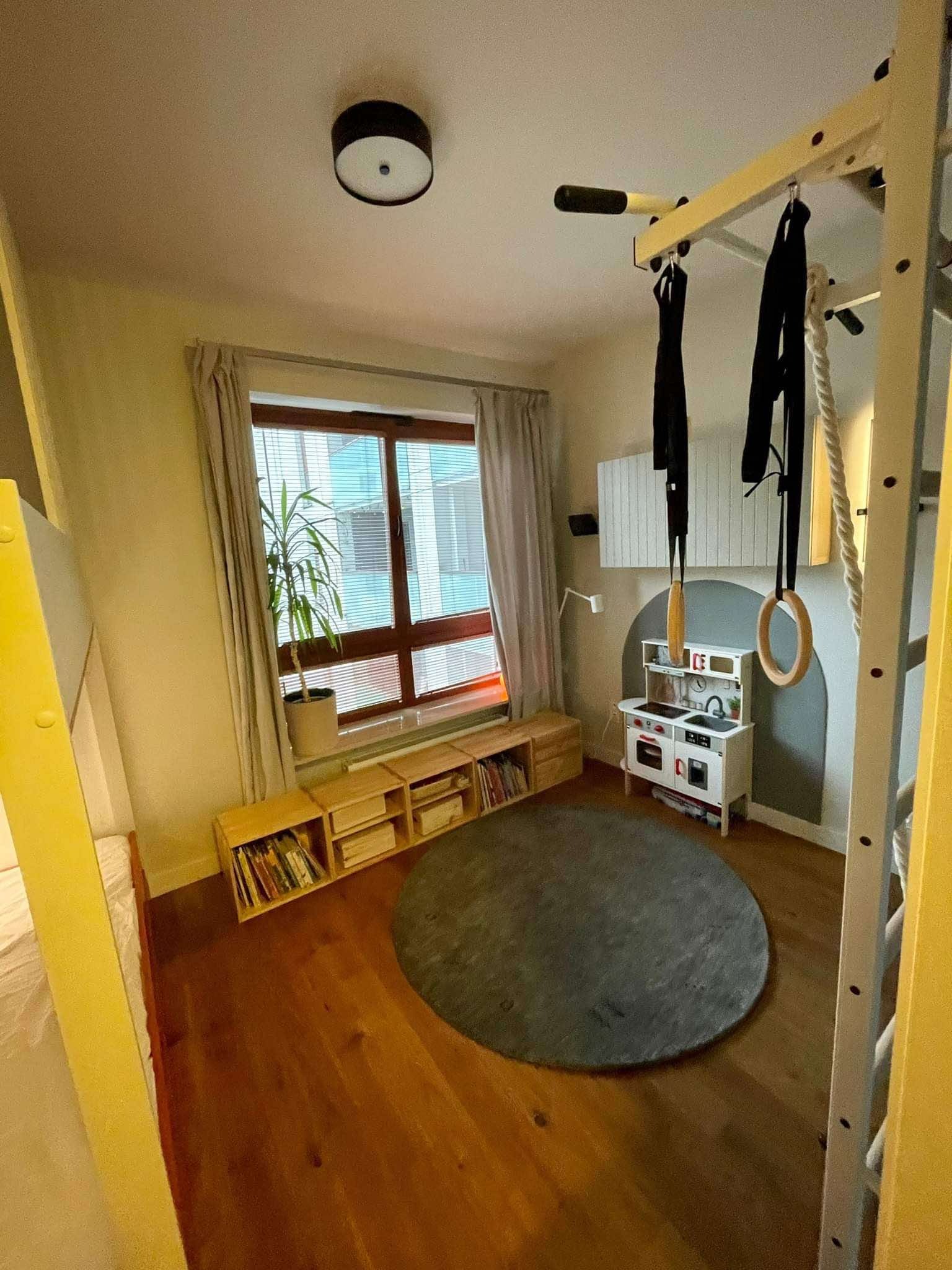 Pokój dziecięcy z drewnianą podłogą, drabinką gimnastyczną, zabawkami i rośliną w doniczce przy oknie. Ustawne meble i stonowana kolorystyka.