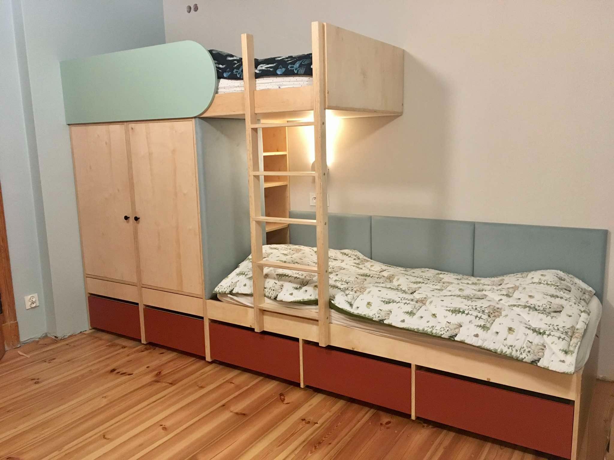 Drewniane łóżko piętrowe z szafą i szufladami w pokoju dziecięcym. Naturalne drewno, pastelowe akcenty kolorystyczne. Widoczne drewniane podłogi.