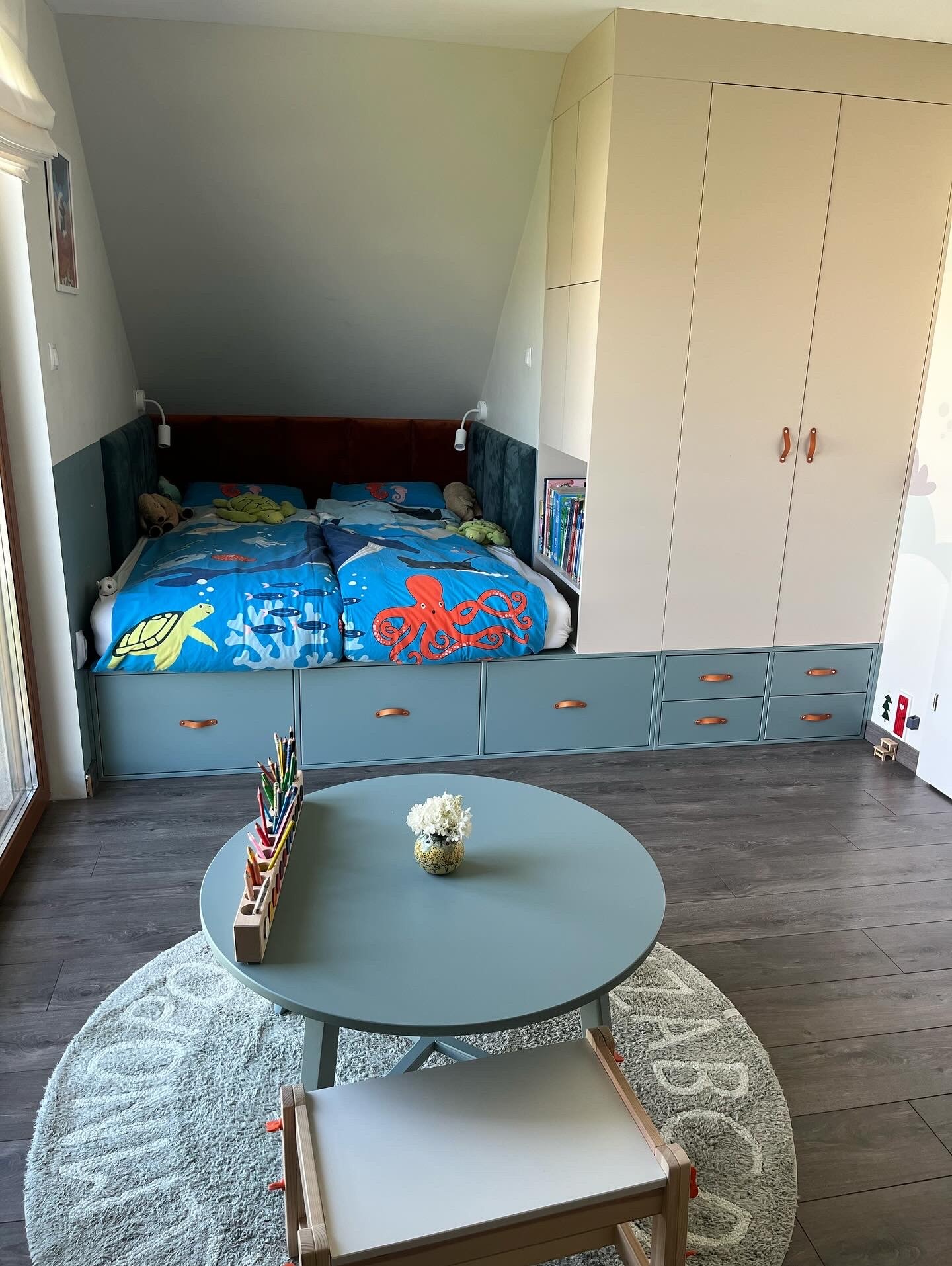 Pokój dziecięcy na poddaszu: łóżko z szufladami w odcieniach błękitu, szafa w zabudowie, okrągły stolik z kredkami i dywan z literami. Przytulne i funkcjonalne wnętrze.