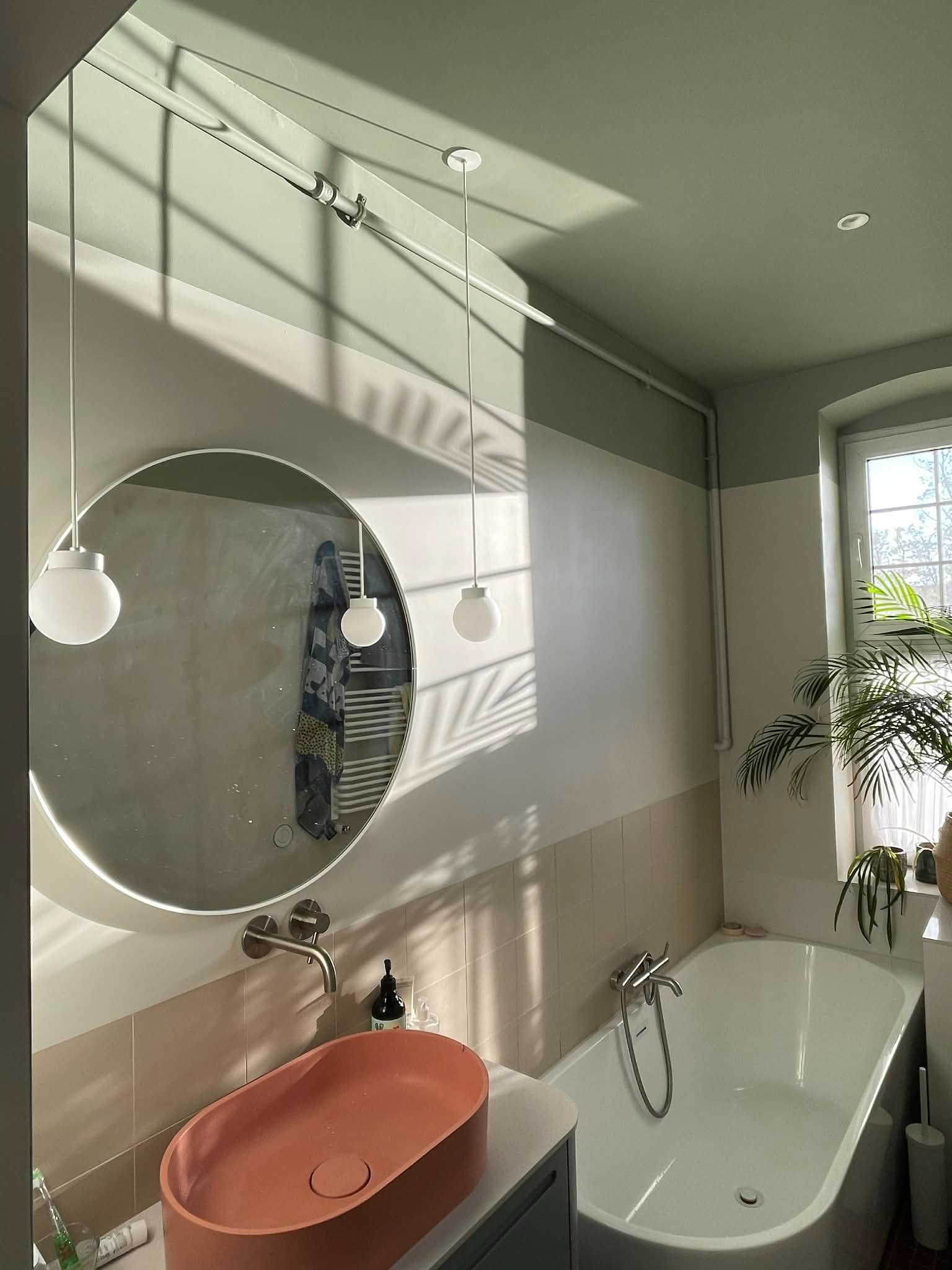 Jasna łazienka z okrągłym lustrem, terakotową umywalką i rośliną w oknie. Ściany w odcieniach zieleni i beżu, z cieniami liści. Minimalistyczny design.