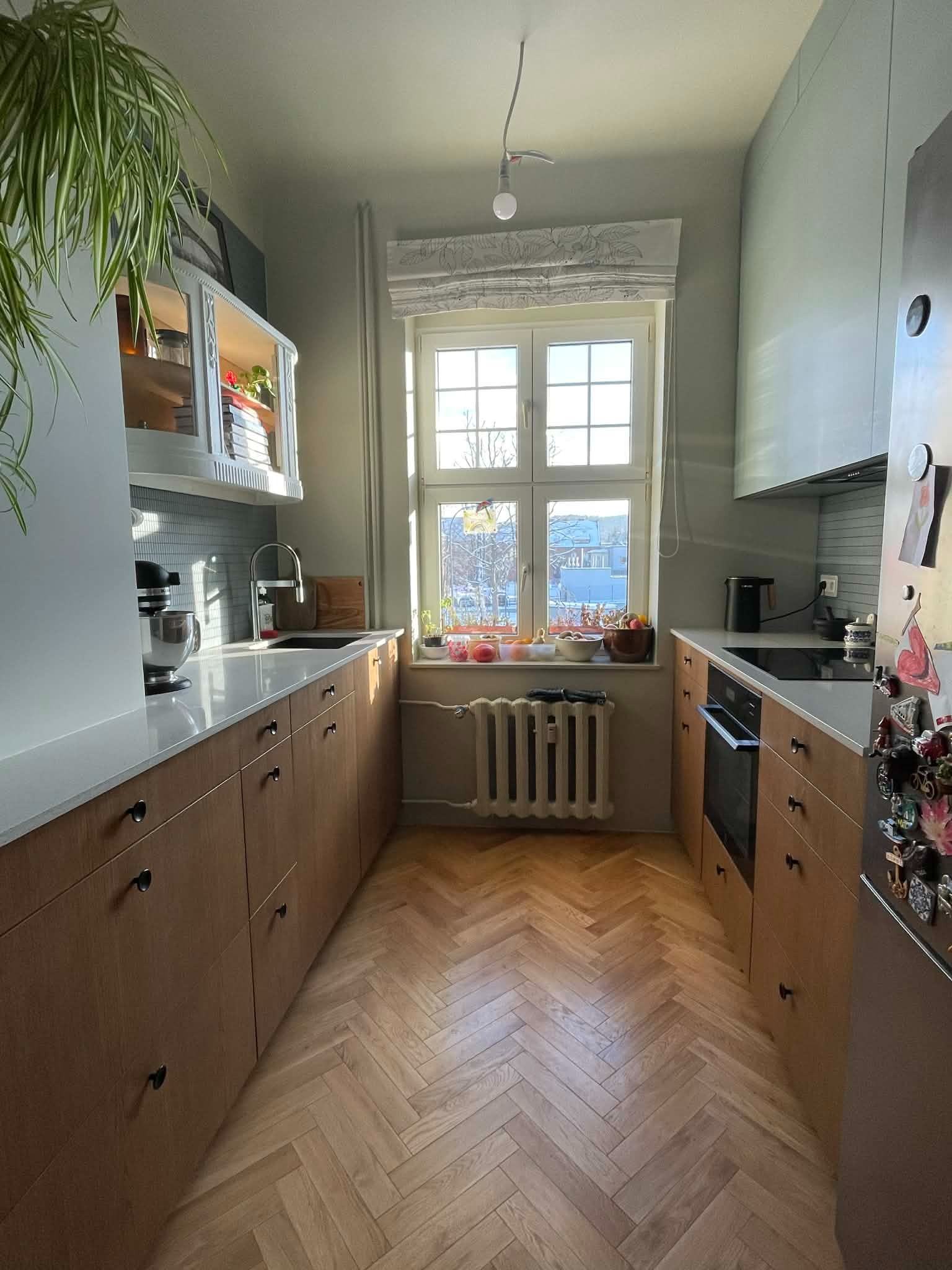 Jasna kuchnia z drewnianymi szafkami, parkietem w jodełkę i dużym oknem z widokiem na miasto. Na blacie mikser i owoce, a na lodówce magnesy.