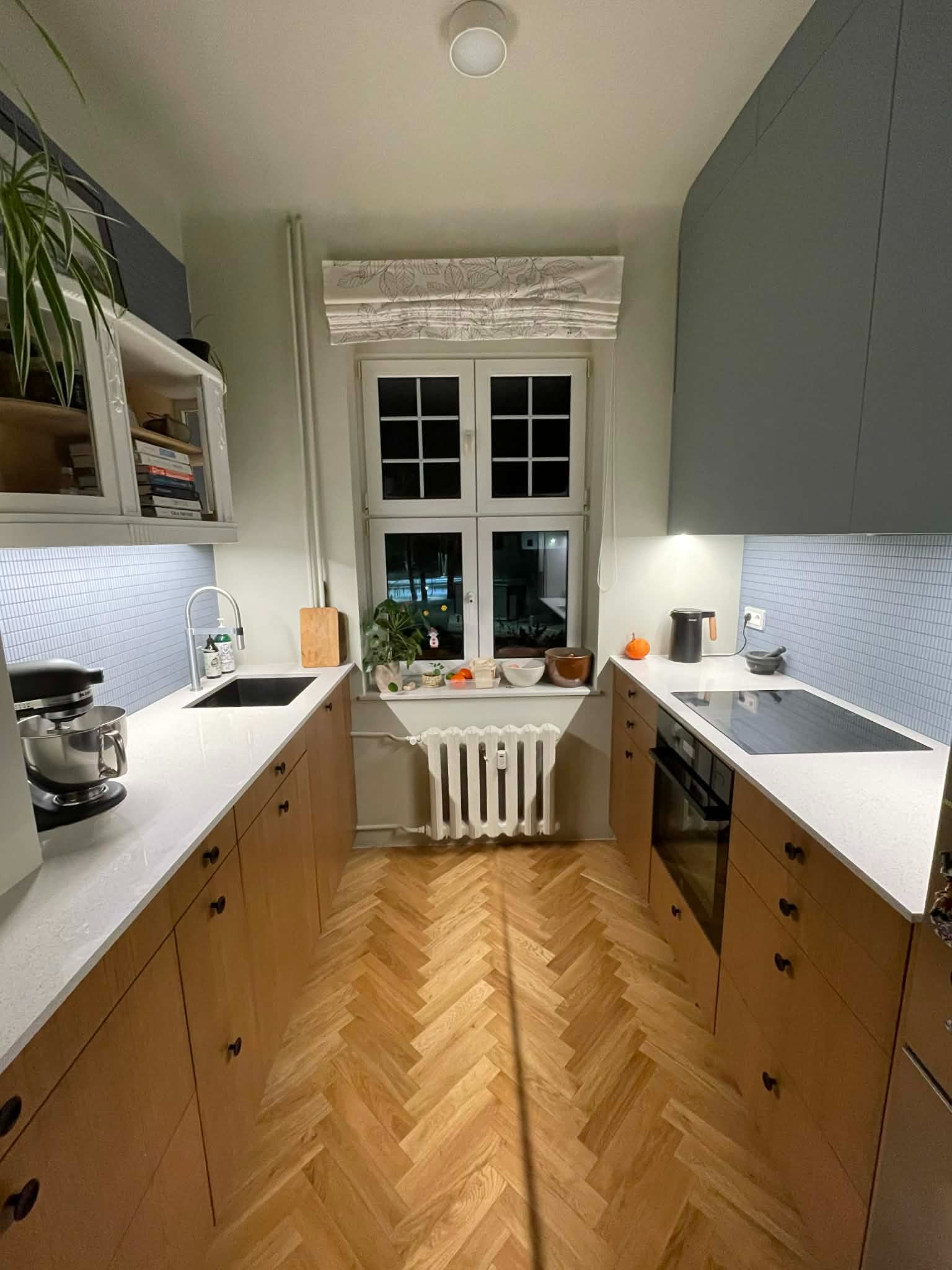 Ustawna kuchnia z drewnianą podłogą w jodełkę, jasnymi blatami i niebieską mozaiką na ścianie. Okno z roletą rzymską i roślinami dodaje przytulności.