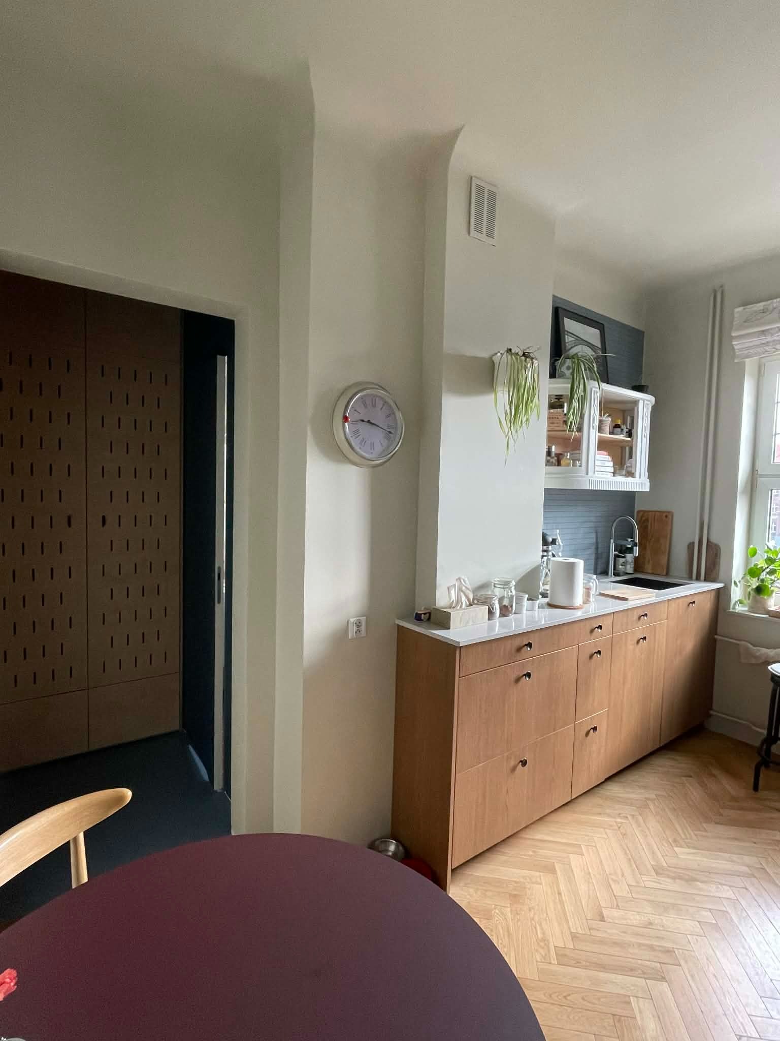 Jasna kuchnia z drewnianymi szafkami, parkietem w jodełkę i okrągłym stołem. Zegar na ścianie. Minimalistyczny design z roślinami doniczkowymi.