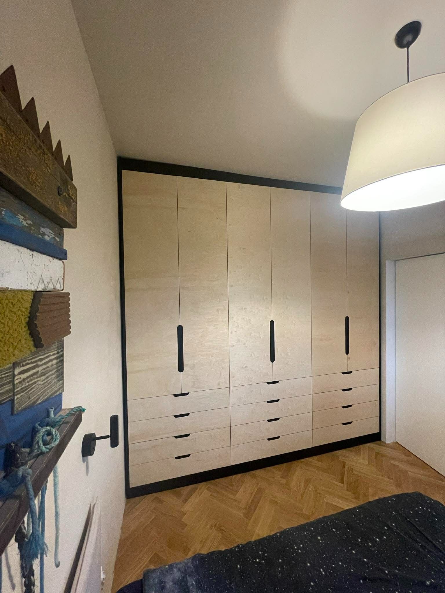 Jasna, drewniana szafa wnękowa z czarnymi detalami w sypialni z parkietem i dekoracjami ściennymi. Minimalistyczny design, ciepłe oświetlenie.