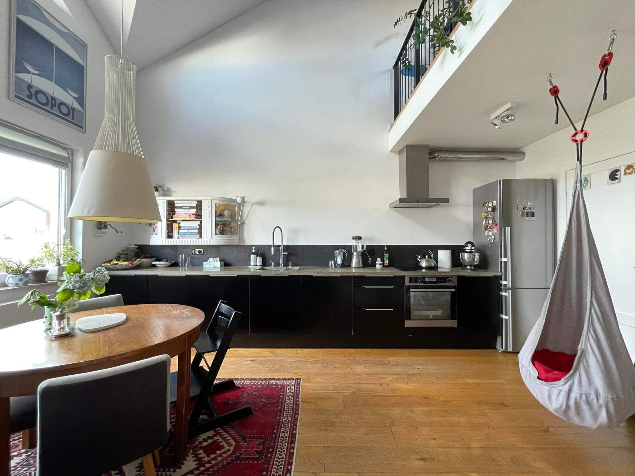 Nowoczesna kuchnia z czarnymi szafkami, drewnianym stołem i wiszącym fotelem hamakowym. Plakat Sopot na ścianie, antresola z roślinami. Jasne drewno na podłodze.