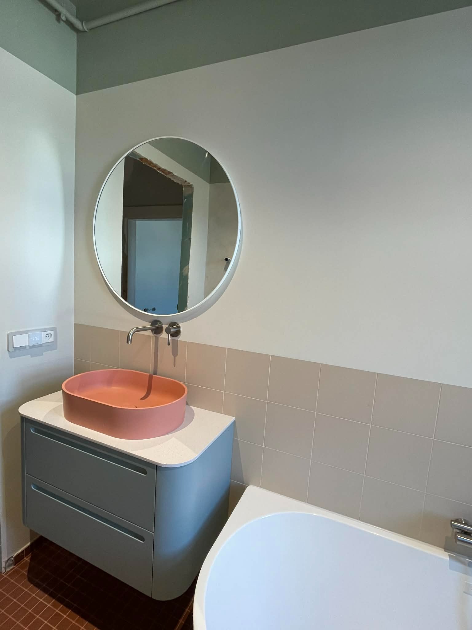 Nowoczesna łazienka z okrągłym lustrem, umywalką w kolorze koralowym i szarą szafką. Widoczne płytki w odcieniach beżu i fragment wanny. Minimalistyczny design.