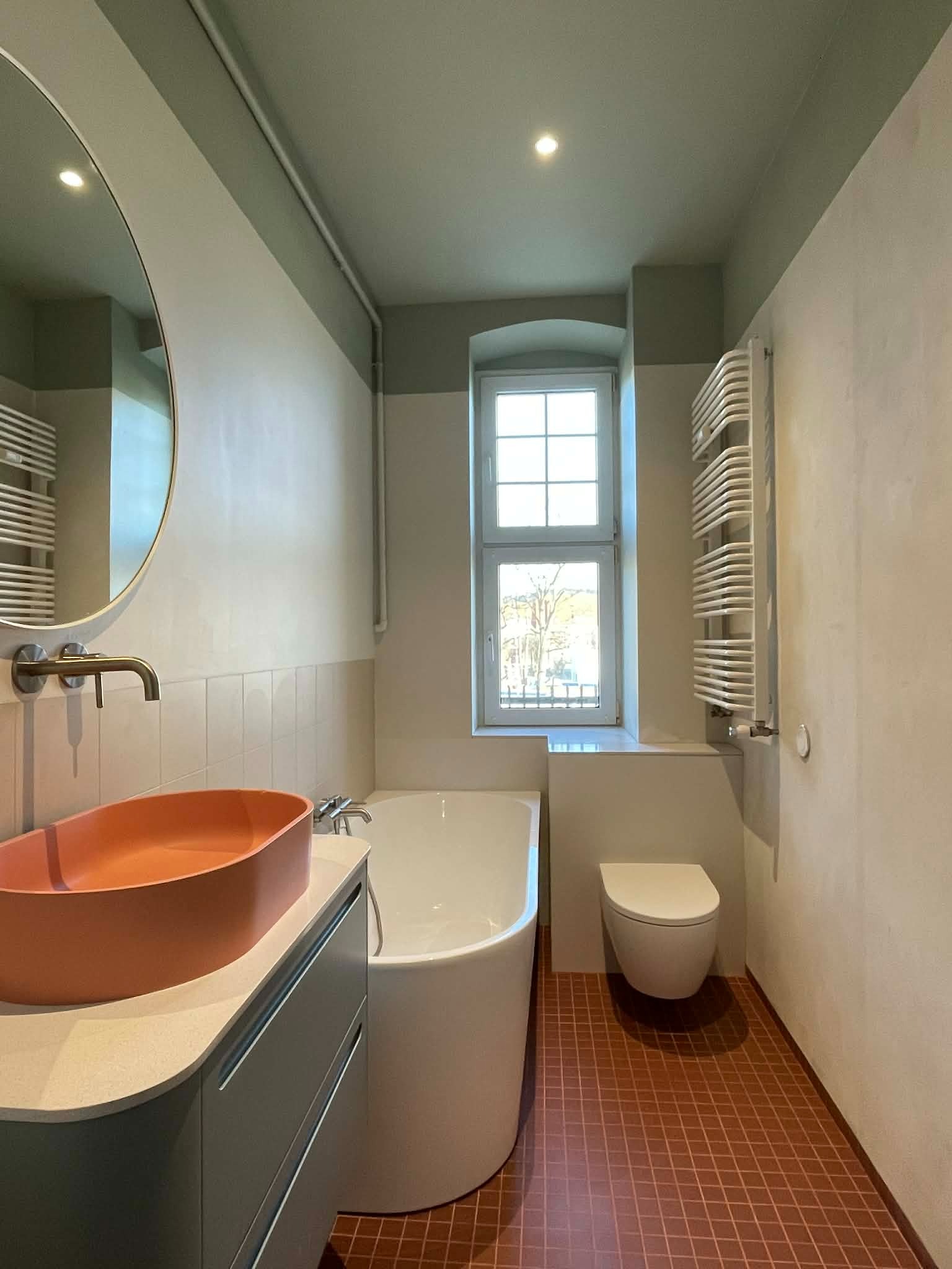 Nowoczesna łazienka z pomarańczową umywalką, białą wanną i grzejnikiem drabinkowym. Ciemna podłoga z płytek i okno z widokiem na zewnątrz.